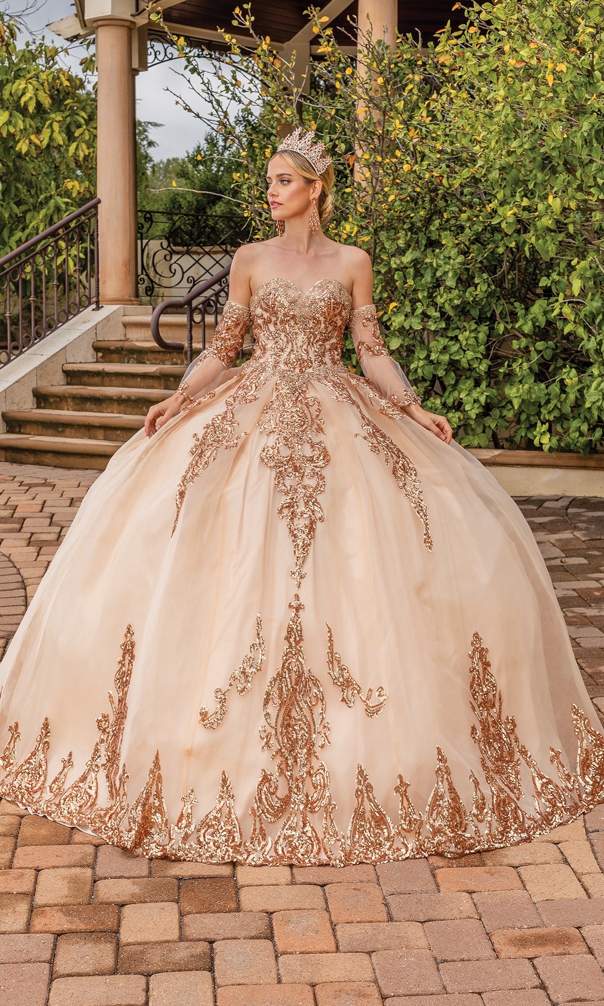 A woman in the Quinceanera Dress 1512 by Dancing Queen, featuring a jeweled crown and ornate gold-embroidered strapless sweetheart bodice, stands outdoors on a stone path amid greenery and columns.