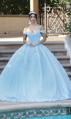 A young woman wearing the Quinceanera Dress 1527 by Dancing Queen—a blue off-the-shoulder ball gown with a sweetheart neckline and tiara—stands by outdoor steps, posing near a pool.