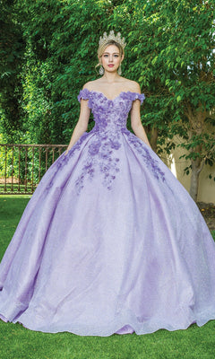 A woman stands outdoors in the Quinceanera Dress 1527 by Dancing Queen, featuring a sparkling lavender ball gown with floral details, an off-the-shoulder neckline, and a tiara.