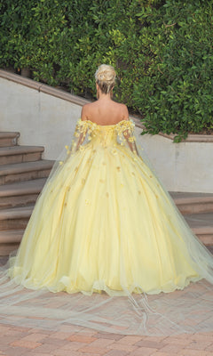 A woman with blonde hair in an updo wears the Quinceanera Dress 1527 by Dancing Queen—a yellow ball gown with floral embellishments, off-the-shoulder neckline, and a long tulle train—while standing on outdoor steps.