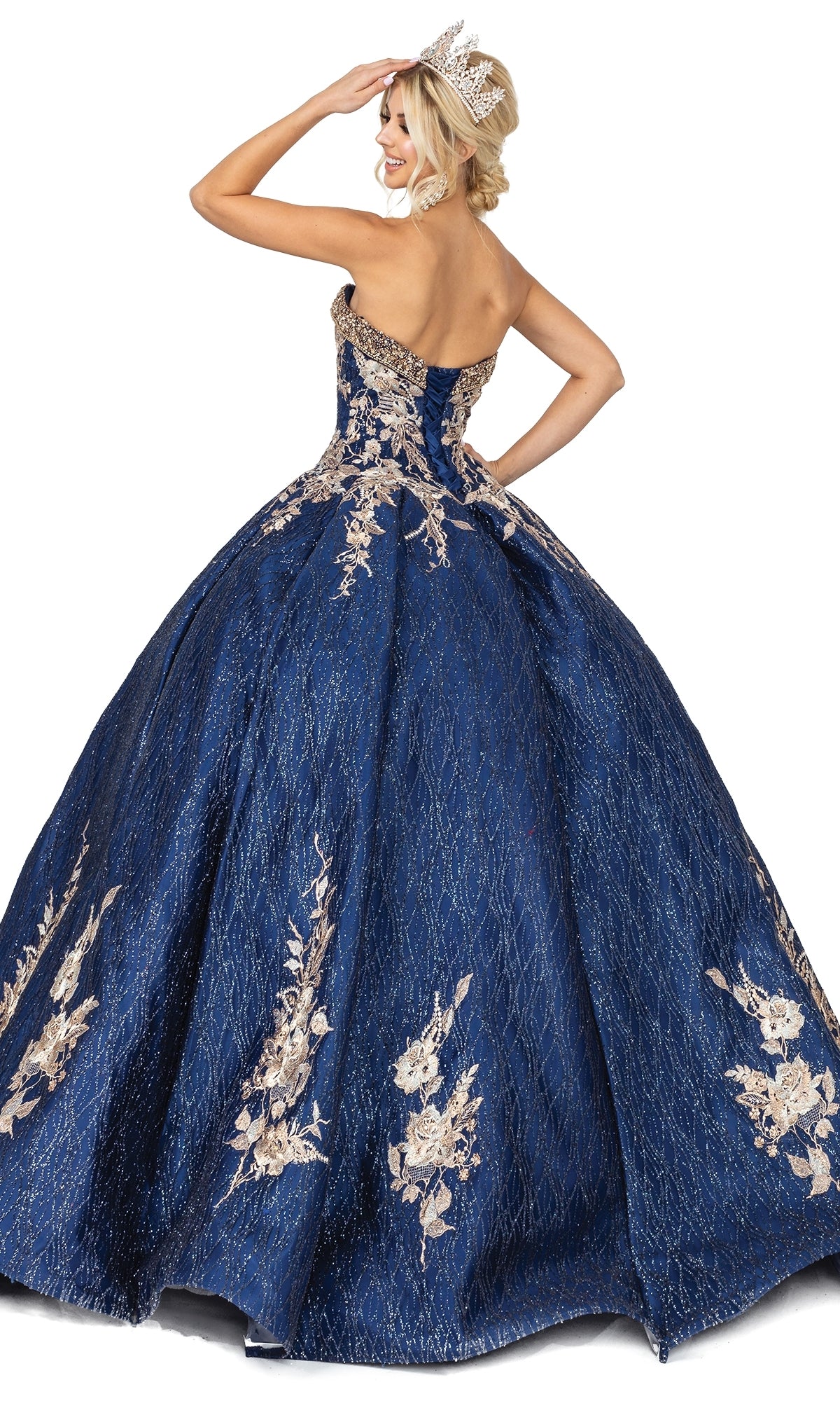 A woman poses facing away, hand raised to her head, wearing Quinceanera Dress 1541 by Dancing Queen—a strapless blue gown with gold floral embroidery and a silver tiara—against a white background.