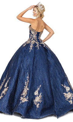 A woman poses facing away, hand raised to her head, wearing Quinceanera Dress 1541 by Dancing Queen—a strapless blue gown with gold floral embroidery and a silver tiara—against a white background.