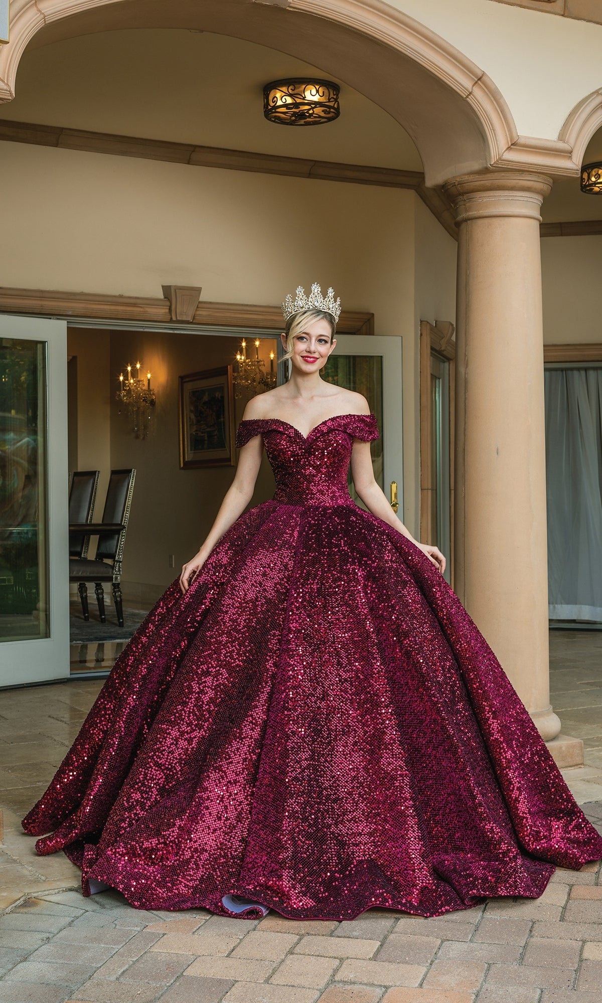 A person wearing the Quinceanera Dress 1559 by Dancing Queen—a sparkling burgundy off-the-shoulder ball gown—and a large crown stands in front of a building with columns and open double doors.