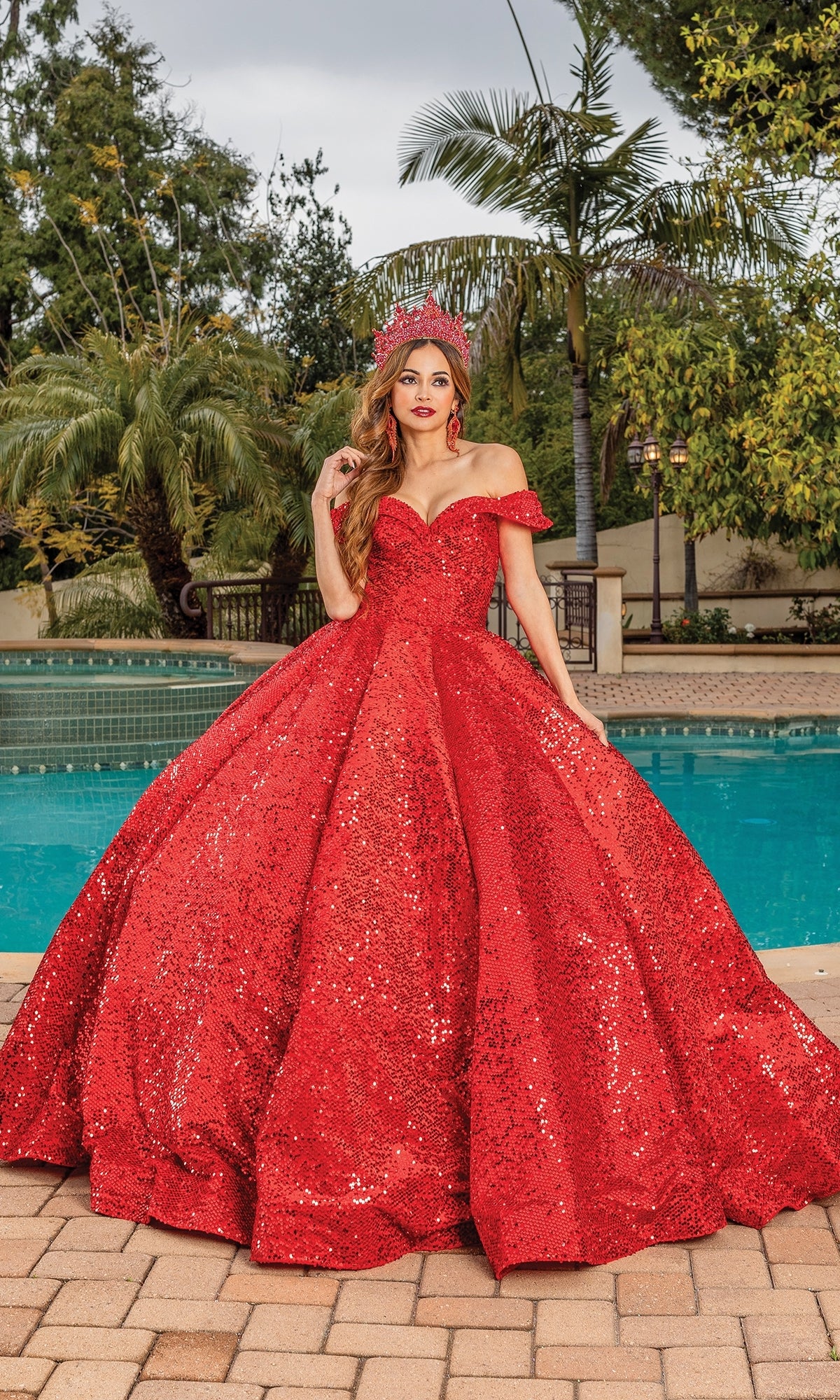 A woman stands by a pool in the Quinceanera Dress 1559 by Dancing Queen and a matching tiara, with palm trees and lush greenery in the background.