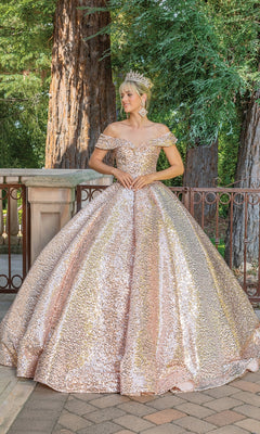 A woman in the off-shoulder, gold and pink sequin Quinceanera Dress 1559 by Dancing Queen with a tiara stands outdoors on a paved area, framed by large trees and a metal railing in the background.