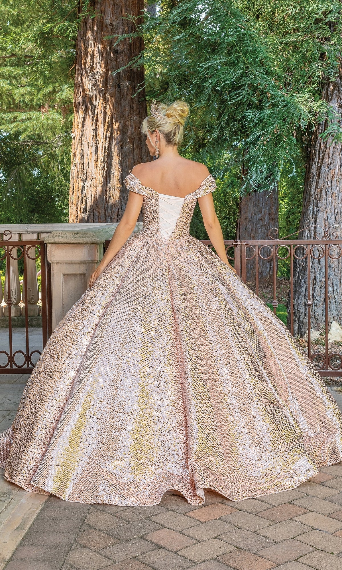 A woman stands outdoors facing away, wearing the Quinceanera Dress 1559 by Dancing Queen—a sparkly pink and gold off-shoulder ball gown with a full skirt. Tall trees and a metal railing are visible in the background.