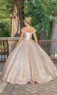 A woman stands outdoors facing away, wearing the Quinceanera Dress 1559 by Dancing Queen—a sparkly pink and gold off-shoulder ball gown with a full skirt. Tall trees and a metal railing are visible in the background.