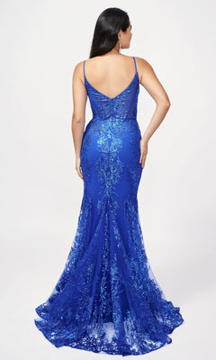 A woman with long dark hair models the Nina Canacci 1562, a blue, floor-length mermaid formal dress with a deep V-back, sequin details, and hot stone accents. She stands against a plain white background.