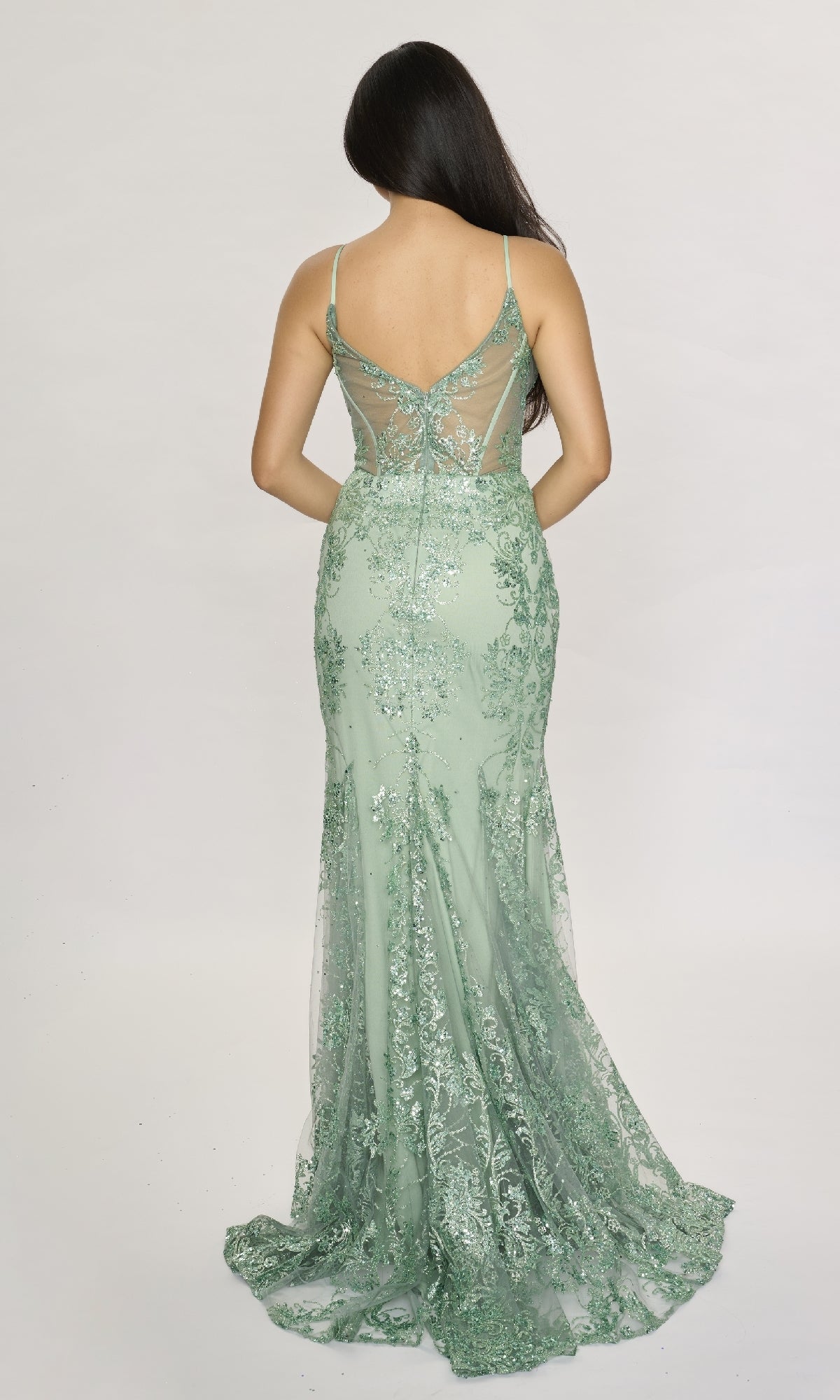 A person stands with their back to the camera, wearing the Nina Canacci 1562—a light green, floor-length mermaid dress featuring lace, sequins, a sheer back panel, and hot stone embellishments for extra sparkle.