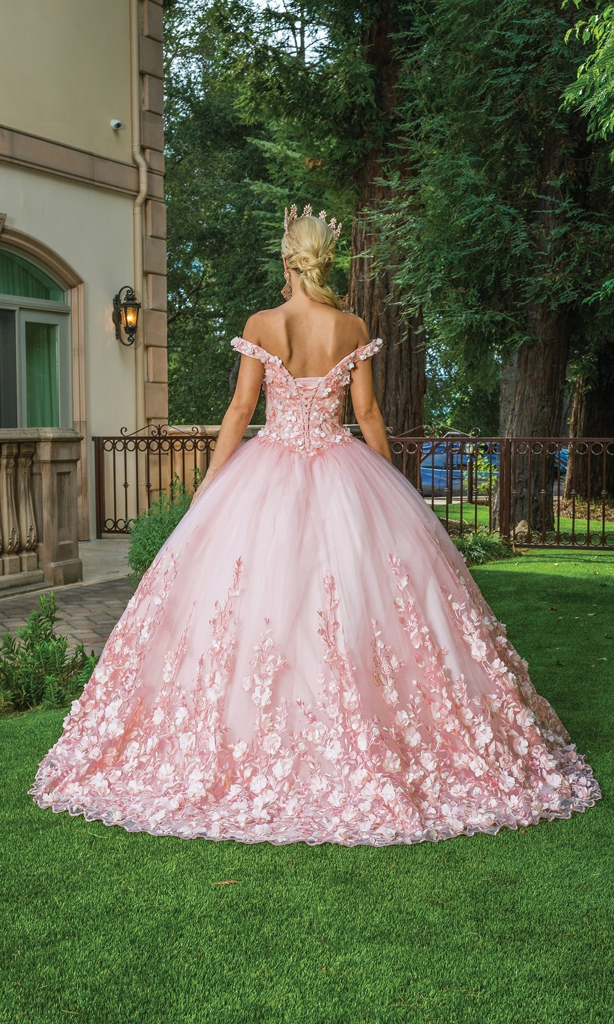 A woman in the Quinceanera Dress 1569 by Dancing Queen, a pink off-the-shoulder ball gown with rose details, stands on grass outdoors facing away near a building and trees.