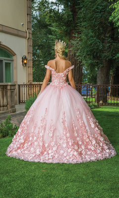 A woman in the Quinceanera Dress 1569 by Dancing Queen, a pink off-the-shoulder ball gown with rose details, stands on grass outdoors facing away near a building and trees.