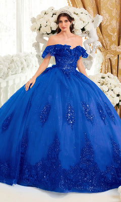 A woman stands indoors wearing the Ladivine 15701 Glitter Quince Dress with Back Bow, a bright blue off-the-shoulder ball gown, surrounded by white flowers and gold curtains.