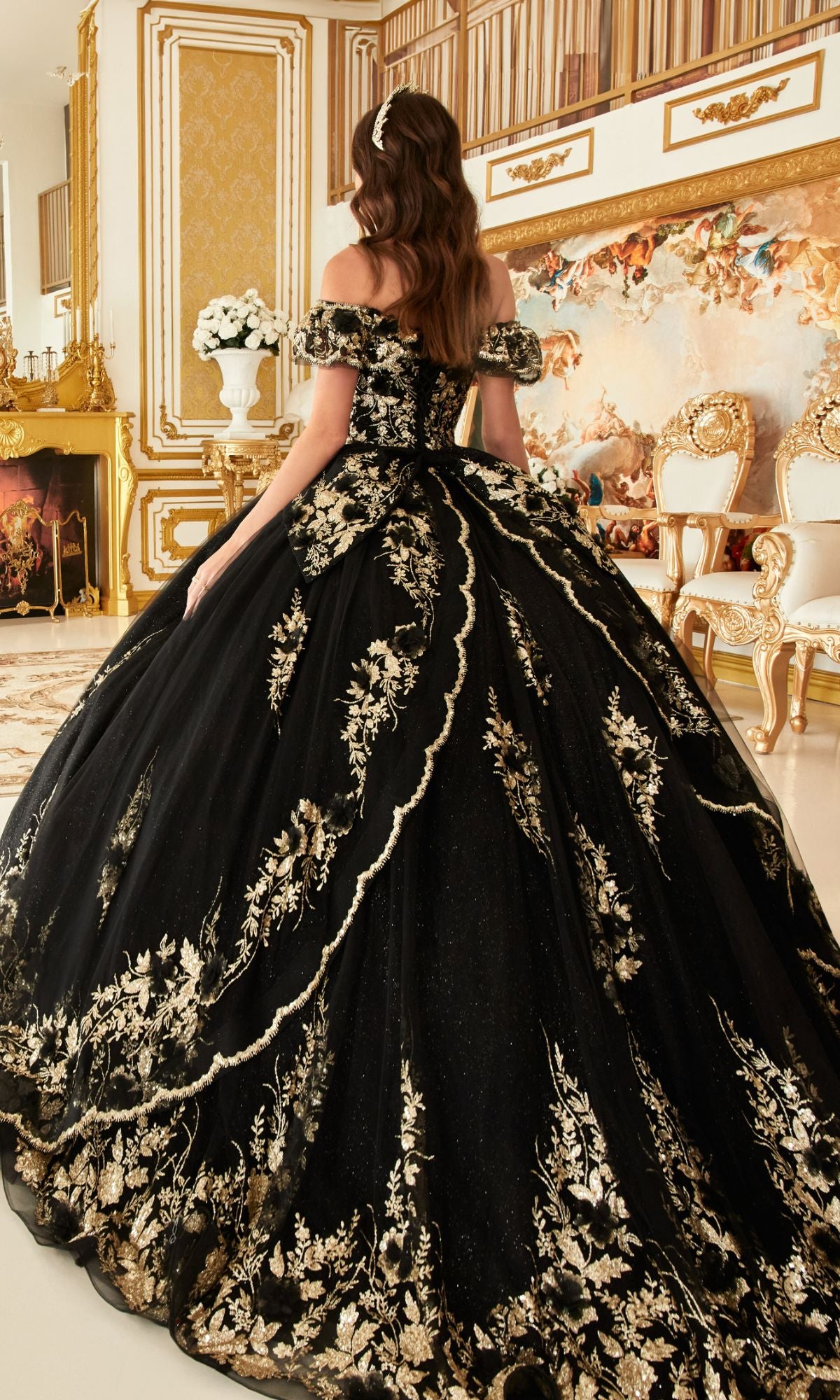 A woman in the Glitter Quince Dress with Back Bow: Ladivine 15701, featuring gold embroidery, stands in an opulent room adorned with gold accents and classical furnishings.