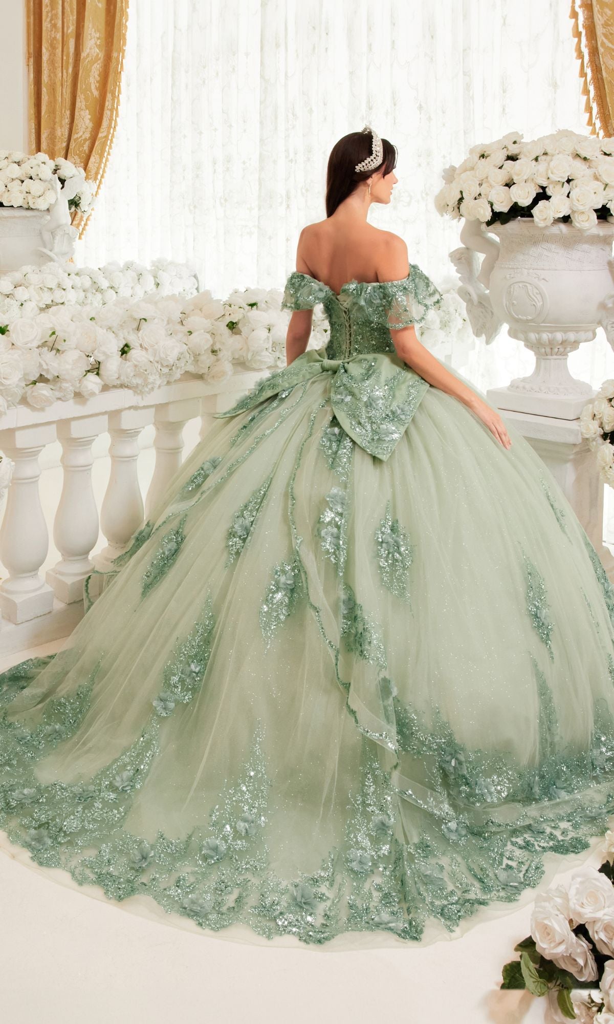 A woman stands with her back to the camera wearing the Ladivine 15701 Glitter Quince Dress with Back Bow—a light green, off-the-shoulder gown with a full sparkling skirt—surrounded by white flowers and ornate decor.