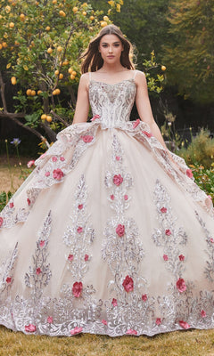 A woman stands outdoors in the Ladivine 15703 Glitter Champagne Quinceañera Dress, featuring silver embroidery and pink floral accents, surrounded by lush greenery and a lemon tree.