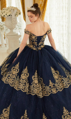 A woman stands indoors, facing away in the Ladivine 15705 Quince Dress with Gold Embroidery—a navy blue off-the-shoulder ball gown. Her hair is in a bun, topped with a crown, radiating elegance.