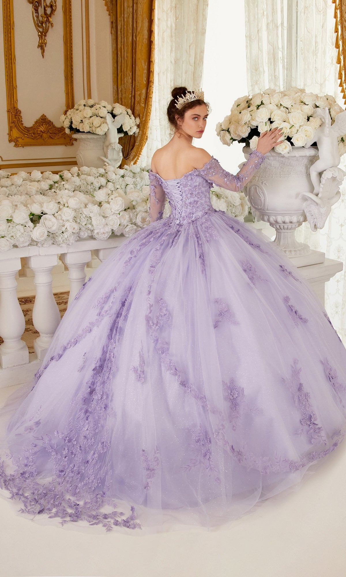 A woman in the Ladivine 15706 Lilac Purple Quinceañera Dress with beaded embroidery and a tiara stands indoors, surrounded by white flower arrangements and ornate decor.