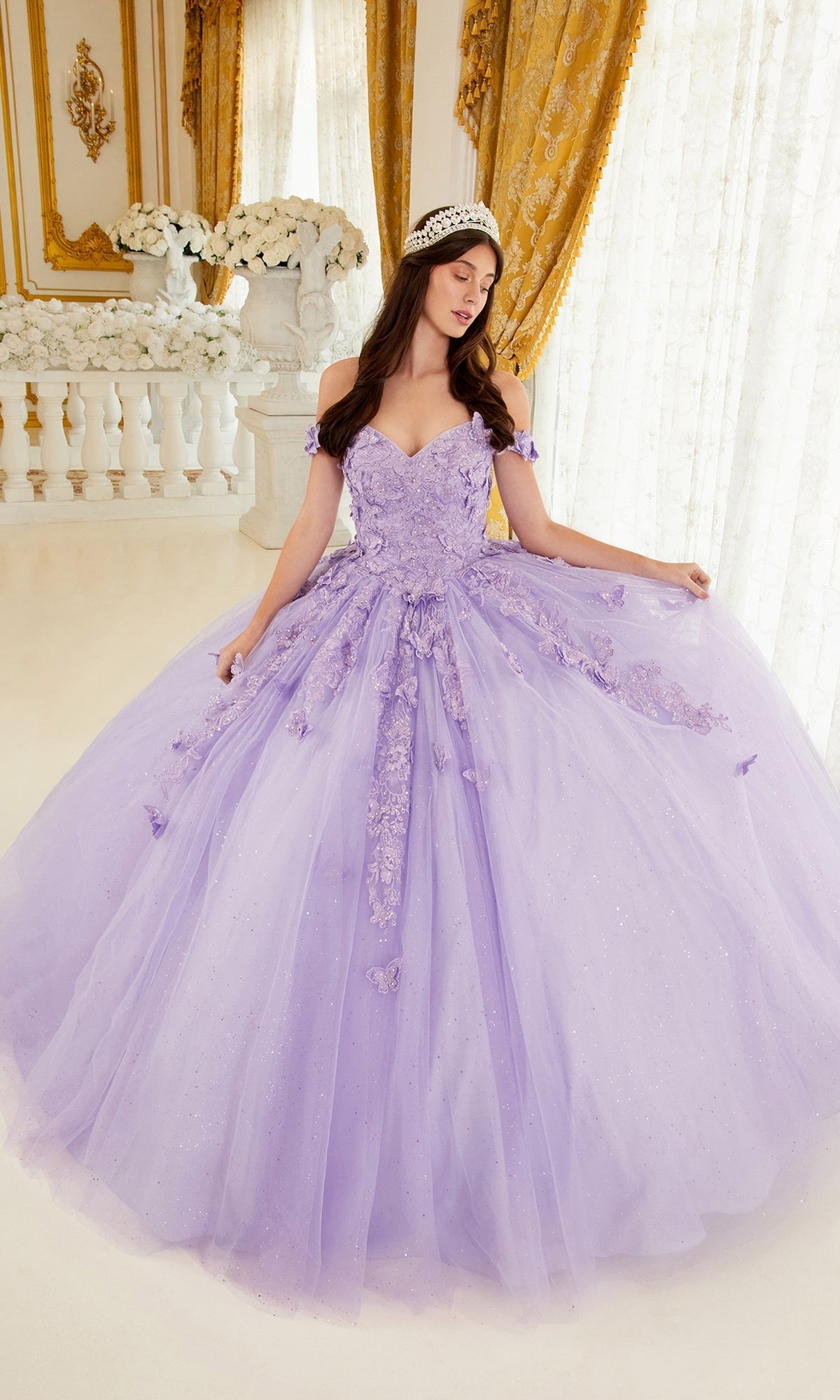 A woman in the Ladivine 15709 Lavender Purple Quinceañera Dress with off-the-shoulder floral embroidery stands indoors beside golden curtains and white flowers.