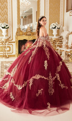A woman wearing the Ladivine 15711 Quinceañera Dress with gold embroidery stands in an ornate room featuring gold decor, a fireplace, and white flower arrangements.