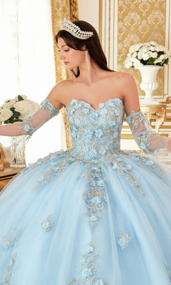 A woman wears the Quinceanera Dress 15714 by Ladivine, a pastel ball gown with lace embroidery and sheer sleeves, paired with a jeweled tiara. She stands indoors, surrounded by white roses and ornate gold décor.