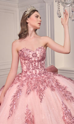 A woman in the Ladivine Strapless Quinceanera Dress 15715, a pink ball gown with sparkling sequins and a tiara, stands indoors and gracefully touches a crystal chandelier.