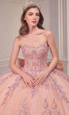 A woman poses indoors against a white paneled wall wearing the Ladivine Strapless Quinceanera Dress 15715, a pink gown featuring an embellished ball gown skirt and paired with a jeweled crown.
