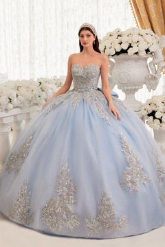 A woman stands indoors among large white floral arrangements, wearing the Ladivine Strapless Quinceanera Dress 15715—a light blue ball gown with silver embellishments.