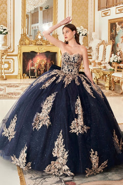 A woman models the Ladivine Strapless Quinceanera Dress 15715—a luxurious gold and navy ball gown—in an ornate, gold-accented room with a fireplace and elegant decor.