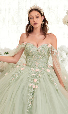 A woman radiates elegance in the Ladivine 15716 Sage Green Quinceañera Dress with pink floral appliqués and a gold crown, posing before a white floral backdrop in her stunning off-shoulder ball gown.