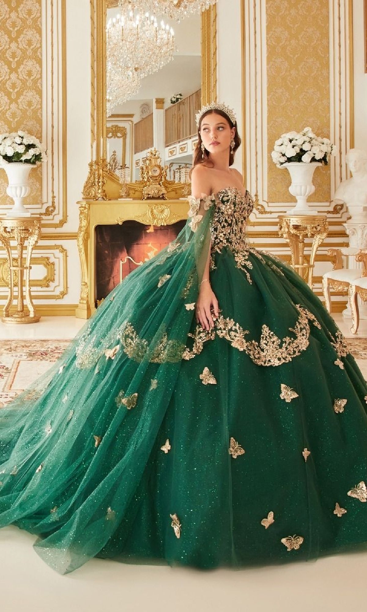 A woman in the Quinceanera Dress 15721 by Ladivine, an emerald green gown with gold embellishments, stands in a luxurious, gold-accented room featuring a fireplace and elegant flower arrangements.