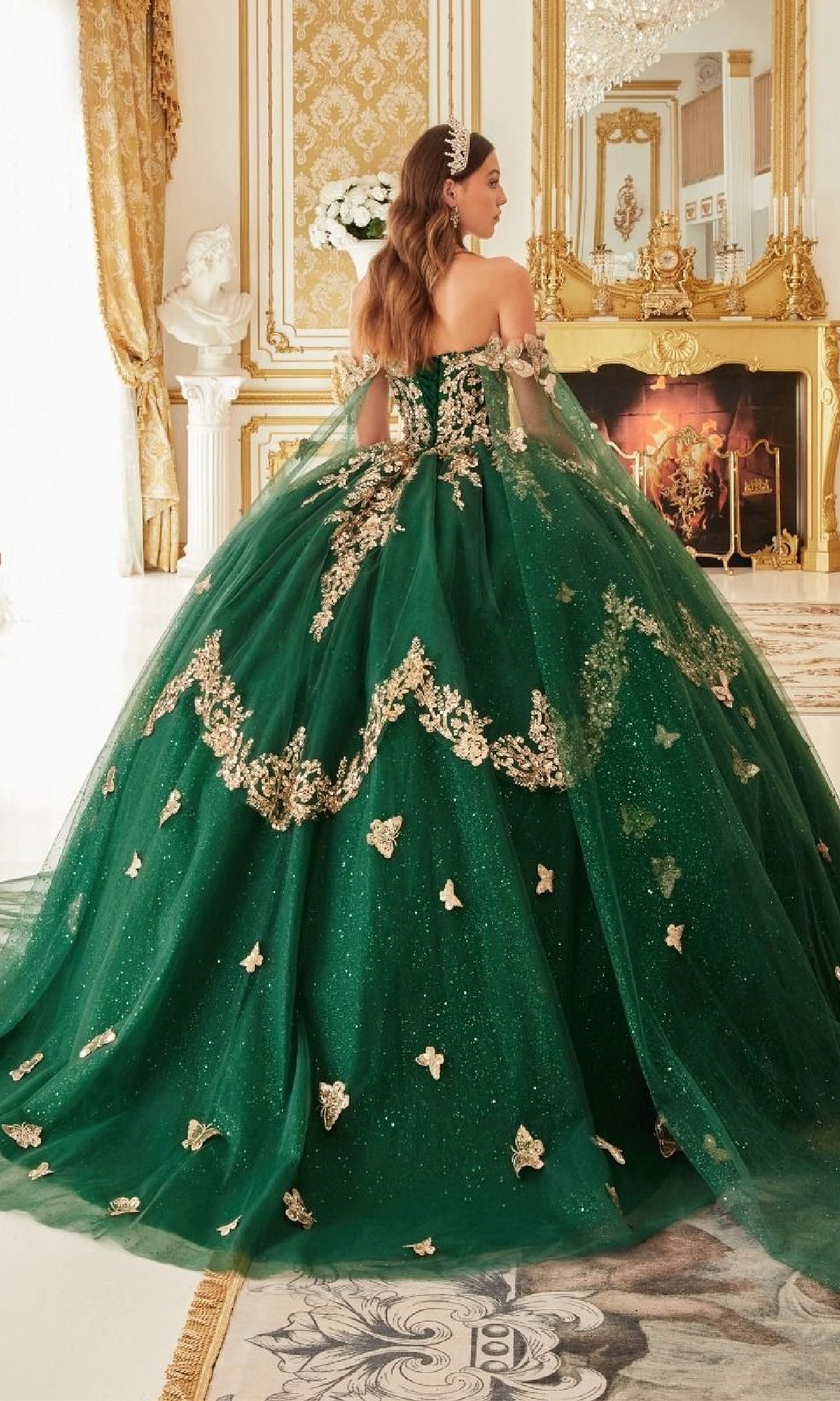 A woman wearing the Quinceanera Dress 15721 by Ladivine stands in an ornate room with gold accents, a fireplace, and a marble statue, her gown shimmering elegantly.