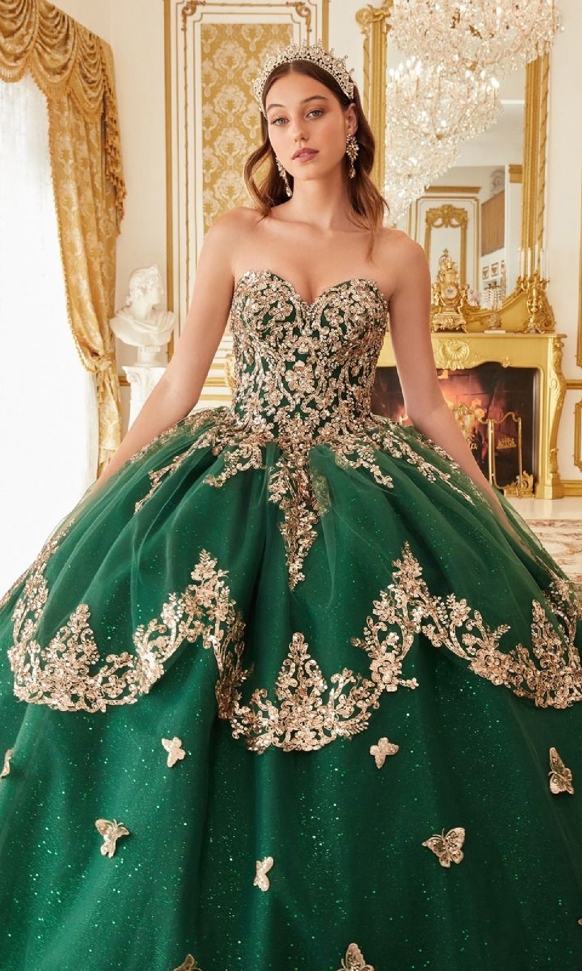 A woman in the Ladivine Quinceanera Dress 15721, an emerald green gown with gold embroidery and a tiara, stands in an ornate room with gold accents, a fireplace, and a chandelier.