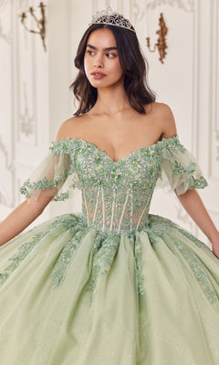 A woman in the Quinceanera Dress 15725 by Ladivine, a sage green off-the-shoulder gown with floral lace appliques and a tiara, stands gracefully in an ornate room, adding elegance to the regal setting.