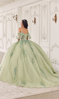 A woman in the Quinceanera Dress 15725 by Ladivine, featuring sage green floral lace appliques, stands in an ornate white room with gold wall sconces, facing away from the camera.