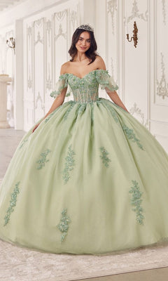 A young woman wearing Quinceanera Dress 15725 by Ladivine in sage green, with off-the-shoulder floral lace appliques and a tiara, stands gracefully in an ornate white room.