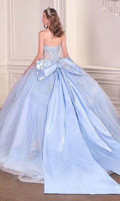 A woman stands in an elegant room, facing away in the Quinceanera Dress 15726 By Ladivine—a strapless, light blue ball gown featuring a large bow, glitter tulle, and a long train.
