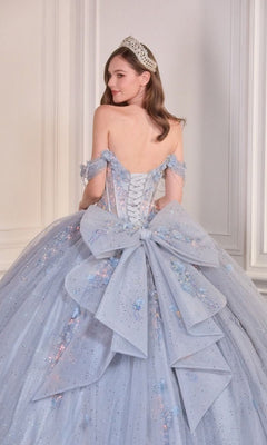 A woman in the Quinceanera Dress 15728 By Ladivine, featuring sparkle tulle, a large back bow, and a tiara, stands before a white paneled wall, glancing over her shoulder.
