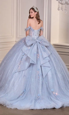 A woman wears the Quinceanera Dress 15728 By Ladivine, a light blue off-the-shoulder ball gown with sparkling details and a large back bow, standing in an elegant room.