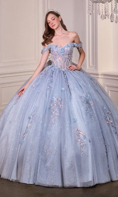 A woman wearing the Quinceanera Dress 15728 By Ladivine, a glitter tulle light blue off-the-shoulder gown, and a tiara stands in an elegant room with white walls.