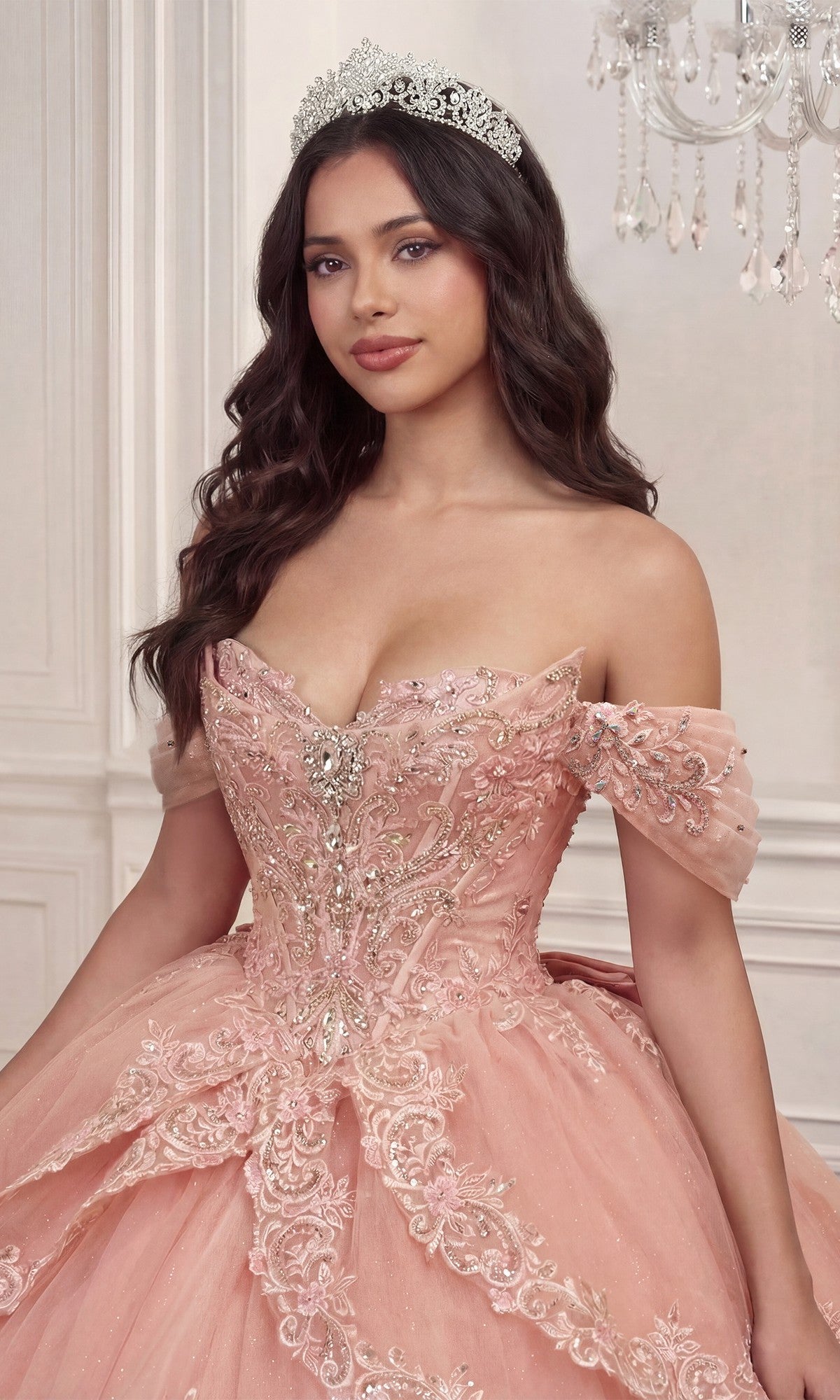 A woman wearing the Quinceanera Dress 15729 by Ladivine—a lavish pink off-the-shoulder ballgown embellished with silver detailing, delicate floral lace appliques, and styled with a sparkling tiara—stands indoors near a chandelier.