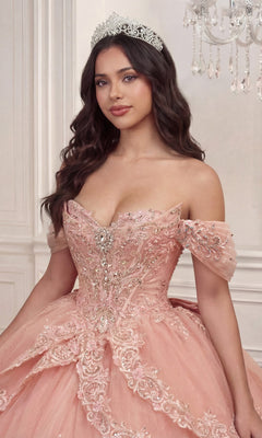 A woman wearing the Quinceanera Dress 15729 by Ladivine—a lavish pink off-the-shoulder ballgown embellished with silver detailing, delicate floral lace appliques, and styled with a sparkling tiara—stands indoors near a chandelier.