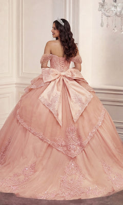 Wearing the Quinceanera Dress 15729 by Ladivine, a woman stands facing away in an ornate room, showcasing the pink off-the-shoulder ballgown embellished with intricate floral lace appliques and a large bow.