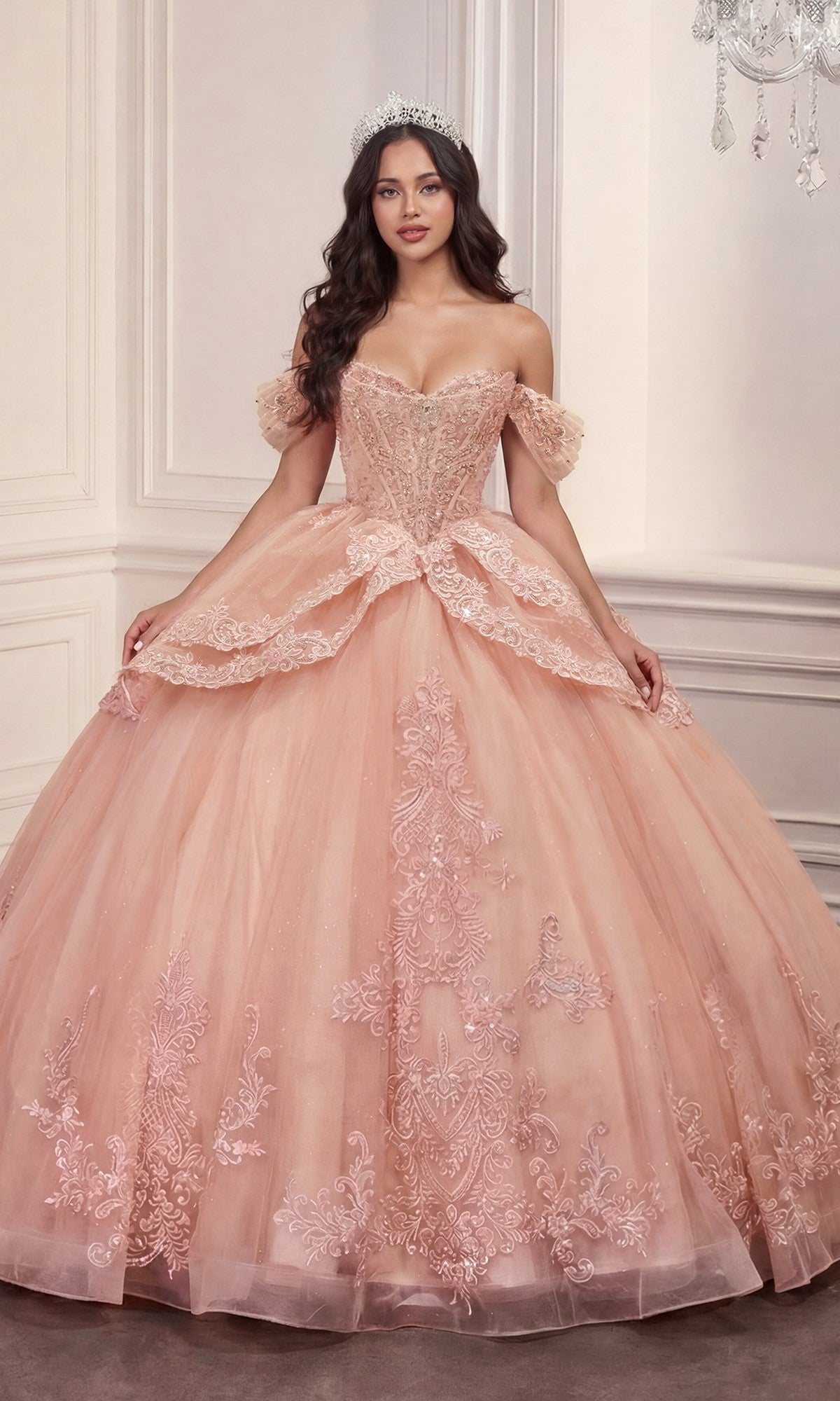 A woman wearing the Quinceanera Dress 15729 By Ladivine in blush, featuring off-the-shoulder floral lace appliques, and a sparkling tiara stands in an elegant, light-colored room.