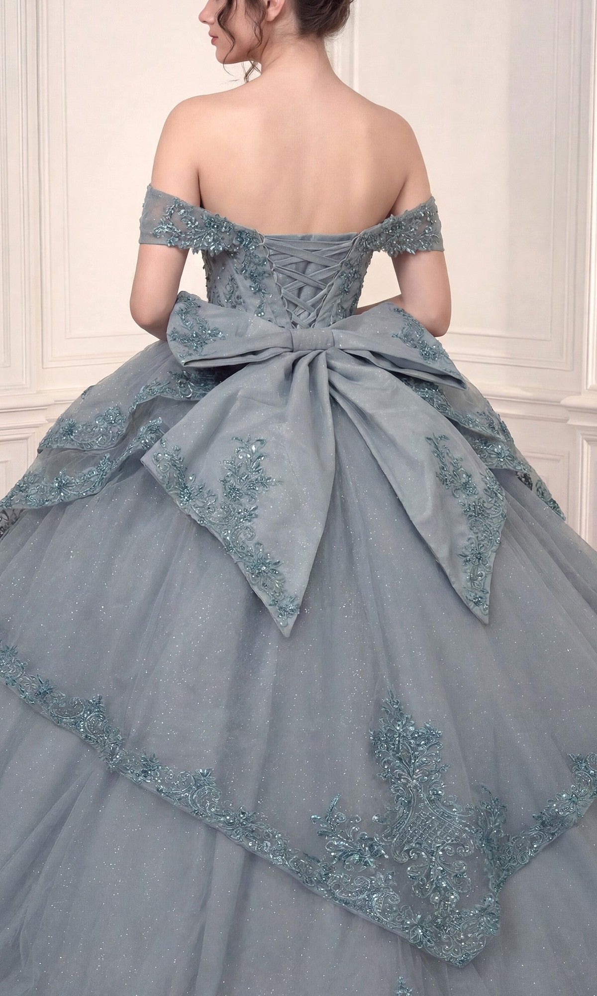 Woman in the Quinceanera Dress 15729 By Ladivine—a blue off-the-shoulder ballgown featuring delicate floral lace appliques and a large bow on the back—standing against a light-colored, paneled wall.