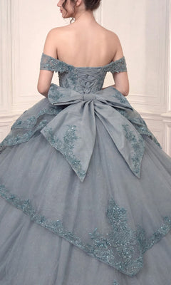 Woman in the Quinceanera Dress 15729 By Ladivine—a blue off-the-shoulder ballgown featuring delicate floral lace appliques and a large bow on the back—standing against a light-colored, paneled wall.