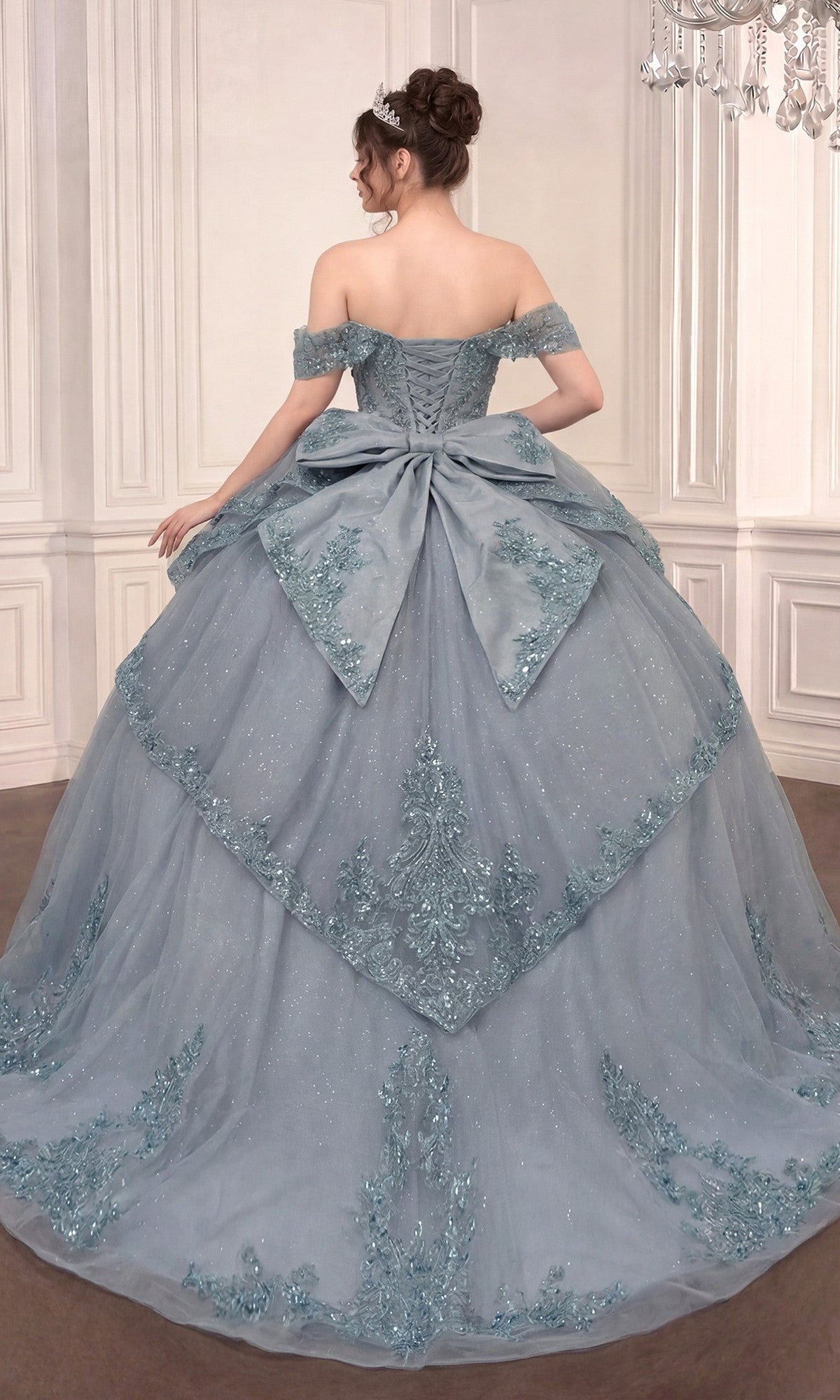 A woman wearing the Quinceanera Dress 15729 by Ladivine—a blue off-the-shoulder ballgown featuring delicate floral lace appliques and a dramatic bow on the back—stands in an ornate room with white paneled walls.
