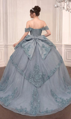 A woman wearing the Quinceanera Dress 15729 by Ladivine—a blue off-the-shoulder ballgown featuring delicate floral lace appliques and a dramatic bow on the back—stands in an ornate room with white paneled walls.
