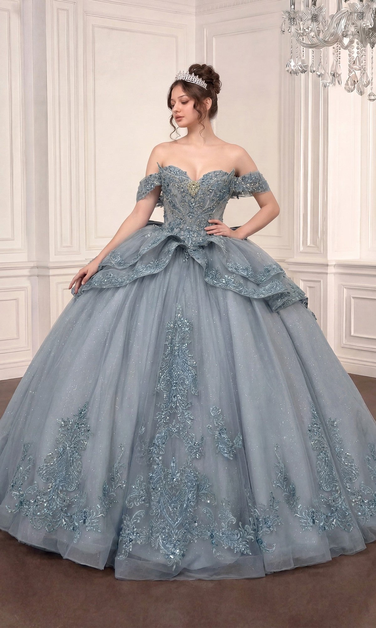 Wearing the Quinceanera Dress 15729 by Ladivine in Dusty Blue, a woman stands in an elegant room with a tiara atop her head, gazing to the side. The ornate off-the-shoulder ballgown features intricate lace and bead details, while delicate floral lace appliques enhance the dress’s regal charm.