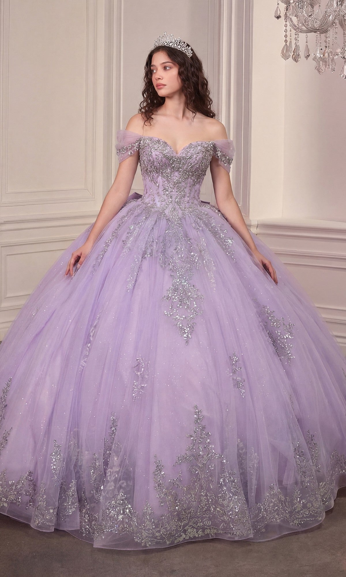 A woman wearing the Quinceanera Dress 15730 by Ladivine in lavender, featuring a crystal-embellished corset bodice and a glitter-tulle ballgown skirt with silver accents and a tiara, stands indoors against an ornate wall.