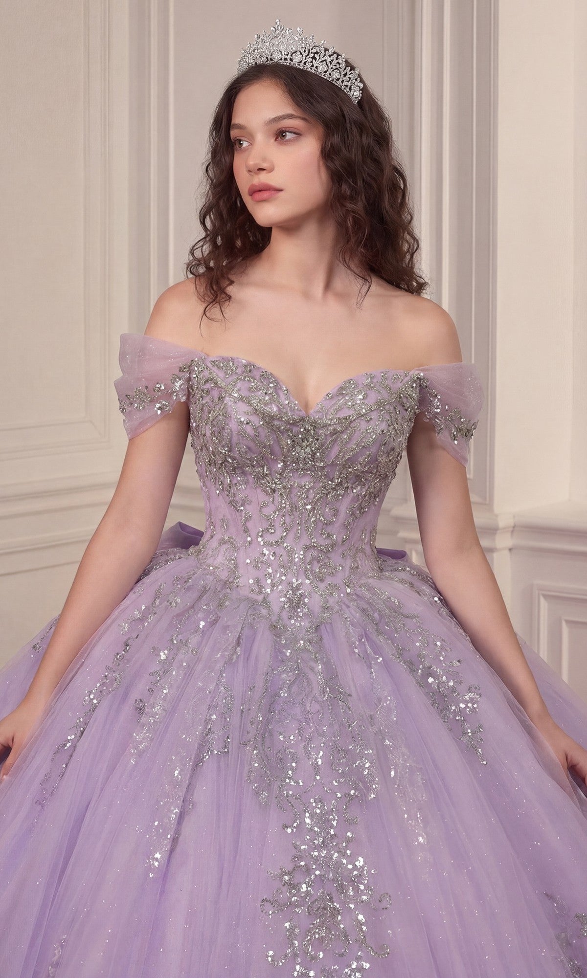 A woman stands in an elegant room with cream-colored walls, wearing the Quinceanera Dress 15730 By Ladivine—a sparkly lavender off-the-shoulder gown featuring a crystal-embellished corset bodice and a glitter-tulle ballgown skirt—her tiara perfectly completing the enchanting look.
